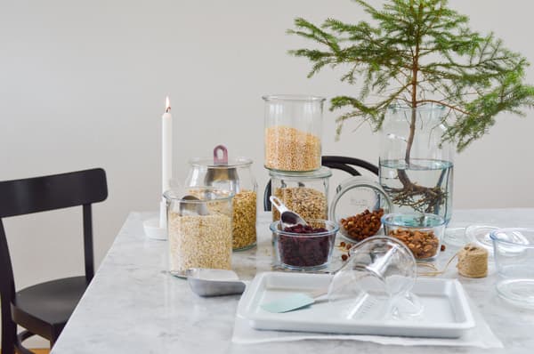 Une scène paisible dans une cuisine, avec des bocaux remplis de céréales et de noix posés sur une table en marbre, une bougie allumée, une petite branche d'épicéa dans l'eau et une chaise noire pliante.