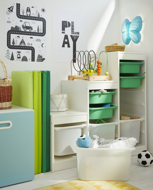 Une structure TROFAST blanche avec bacs de rangement verts et blancs de différentes tailles, un tapis de jeu vert, deux affiches et un tapis jaune au sol.