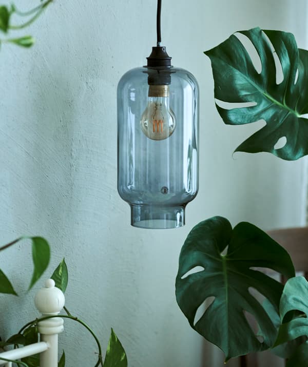 Une suspension en verre bleu suspendue au-dessus d’un cadre de lit blanc à côté d’une plante MONSTERA.