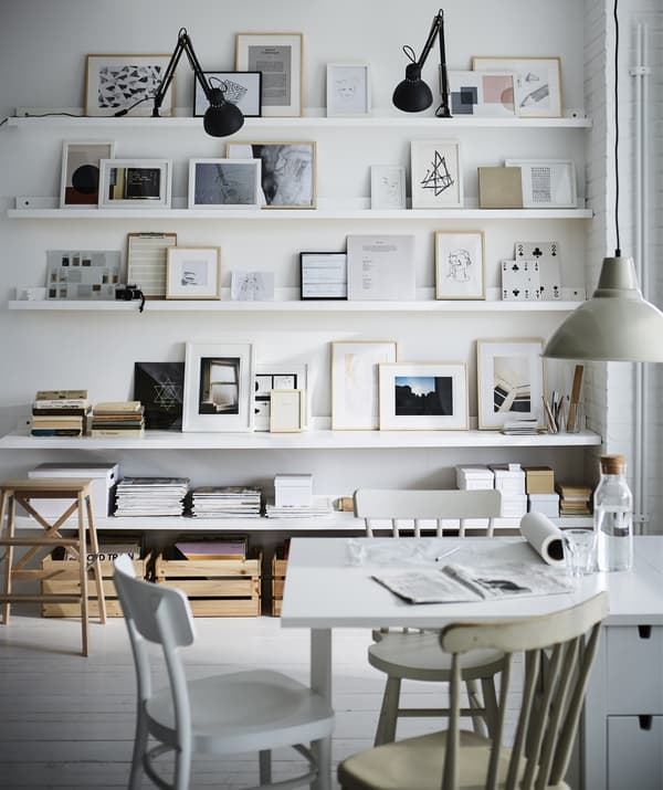 Une table blanche devant une composition de peintures sur des tablettes et des étagères pour photos blanches.