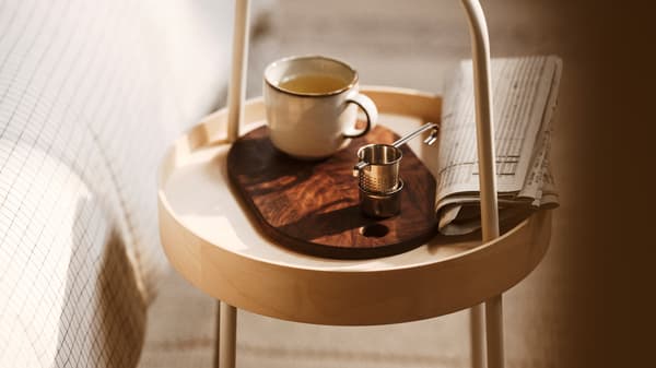 Une table d’appoint BURVIK avec un mug GLADELIG gris posé sur une planche à découper. La table se trouve sur un tapis à poils ras BRÖNDEN.