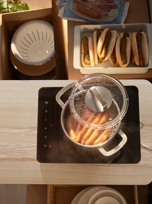 Une table de cuisson à induction TILLREDA sur laquelle des saucisses cuisent dans une casserole d’eau frémissante, à côté d’un plat contenant des hot-dogs végétariens et à la viande.