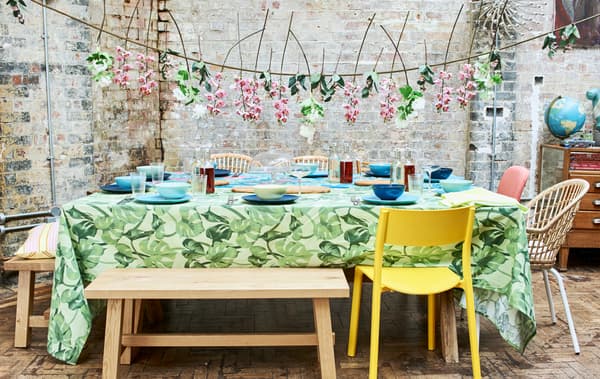 Une table de fête estivale dressée, avec des nappes de couleurs vives, de la vaisselle bleue et verte, des chaises et des bancs et une guirlande de fleurs.