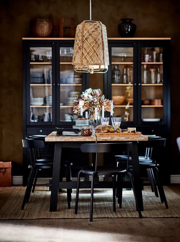 Une table de salle à manger dressée pour un repas de fête, sous une suspension KNIXHULT. Vitrines foncées avec des accessoires de table devant le mur du fond.