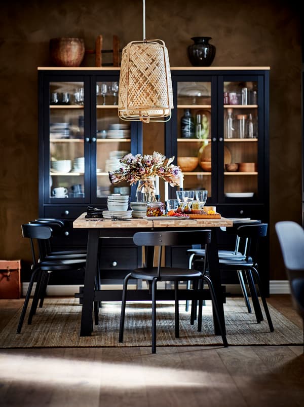 Une table de salle à manger SKOGSTA dressée pour un repas de fête, sous une suspension. Vitrines foncées avec des accessoires de table devant le mur du fond.