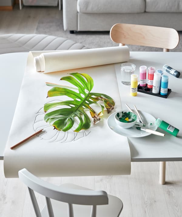 Une table de séjour aménagée pour une session de peinture avec des plantes: de la peinture et du papier à dessiner MÅLA et une feuille un peu flétrie d’une plante MONSTRERA.