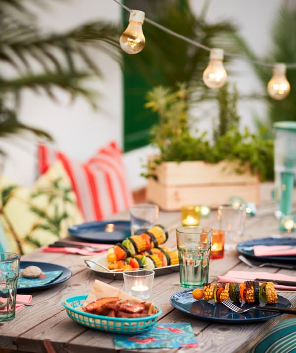 Une table d’extérieur entourée de plantes et de textiles et surmontée d’une guirlande lumineuse, dressée avec des assiettes FÄRGRIK et des couverts LIVNÄRA.
