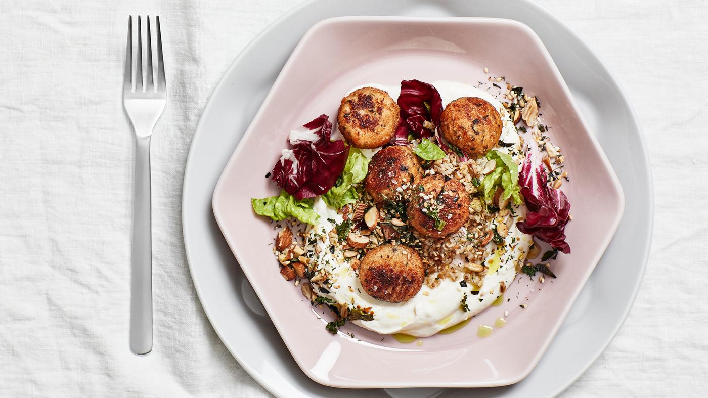 Une table dressée avec une nappe blanche, une fourchette et une assiette de forme hexagonale vieux rose contenant cinq boulettes de cabillaud, de la salade, des graines et du yaourt.