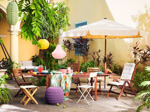Une table en bois et une variété de chaises disposées à l’extérieur contre un mur jaune, avec un parasol et des lanternes colorées.