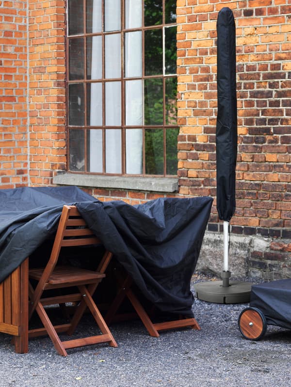 Une table et des chaises d’extérieur en bois, rangées sous une housse noire à côté d’un bâtiment en briques avec une fenêtre et un parasol replié.