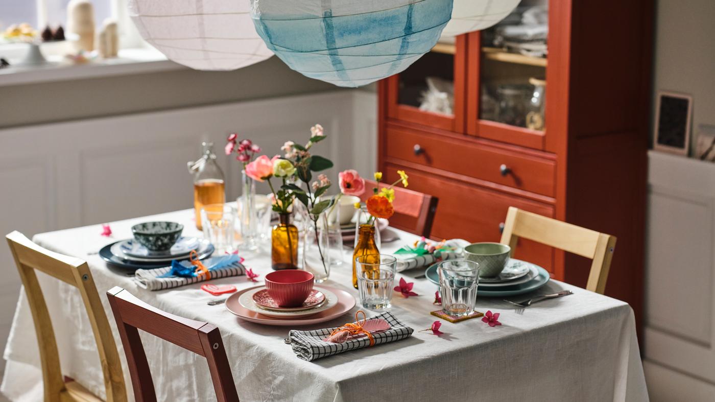 Une table recouverte d’une nappe écrue/beige avec de la vaisselle mélangée de différentes couleurs, des verres POKAL et des fleurs artificielles.