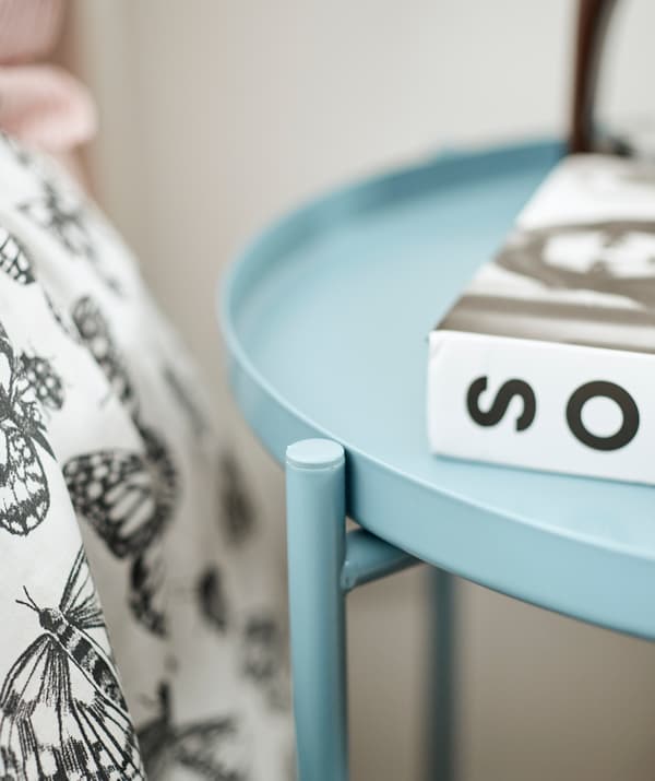 Une table ronde bleue en métal sur laquelle est posé un livre, à côté d’un lit revêtu d’une couette en noir et blanc.