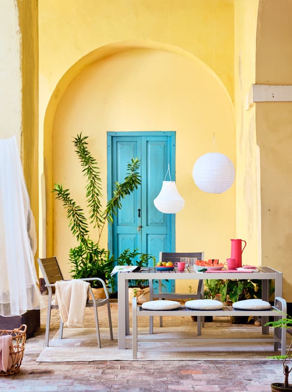 Une table, un banc et des chaises placés sur un tapis, surmontés de lanternes blanches. Porte bleue et mur jaune à l’arrière-plan.