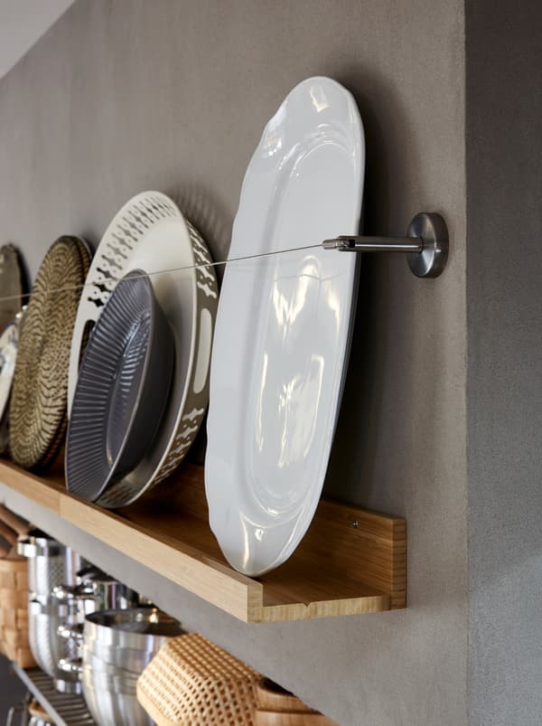 Une tablette pour photos MÅLERÅS en bois mettant en valeur une assiette et d’autres ustensiles de cuisine, avec un câble à rideaux métallique en guise de protection.