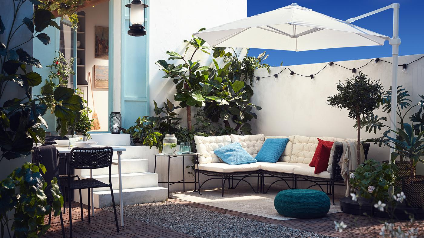 Une terrasse extérieure avec un canapé modulaire, des coussins blancs, un parasol blanc, un pouf bleu-vert et des plantes.