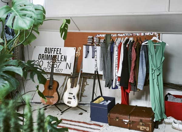 Une tringle remplie de vêtements à côté de deux guitares, une valise et quelques boîtes de rangement.
