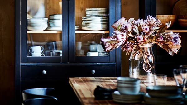 Une vitrine HEMNES noir-brun remplie de verres et d’un service de table, derrière une table de salle à manger ornée d’un vase.