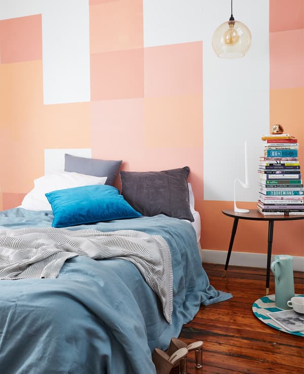 Unha cama con téxtiles azuis e grises en fronte dunha parede pintada con cadros de cor rosa, coral e laranxa.