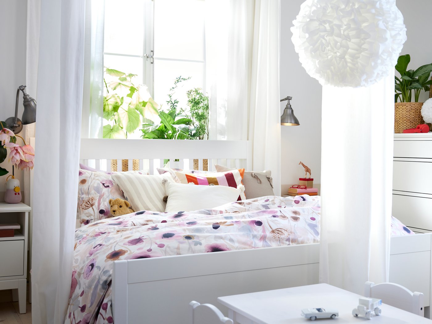 Unha cama IDANÄS branca con roupa de cama LÖNNHÖSTMAL en fronte dunha fiestra. Ao pé da cama hai unha mesa infantil.