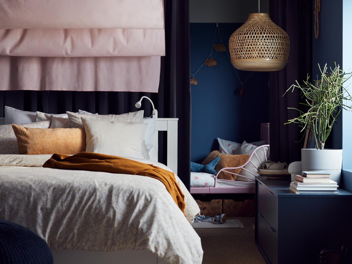 Unha cama SONGESAND branca cuberta con roupa de cama en gris claro cunha manta OMTÄNKSAM amarela enriba e unha cortina rosa detrás.