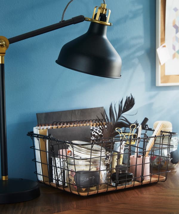 Unha canastra de arame IKEA PLEJA nun escritorio e chea de adornos, cadernos e accesorios de escritorio.