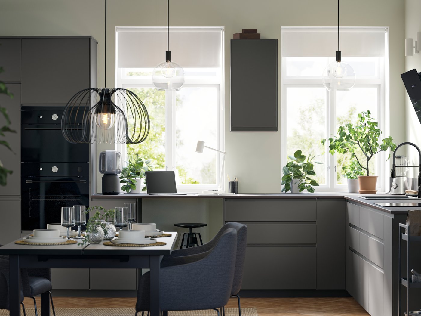 Unha cociña con frontes lisas VOXTORP en gris escuro, con dúas grandes fiestras, unha zona de comedor, unha lámpada colgante e electrodomésticos negros.