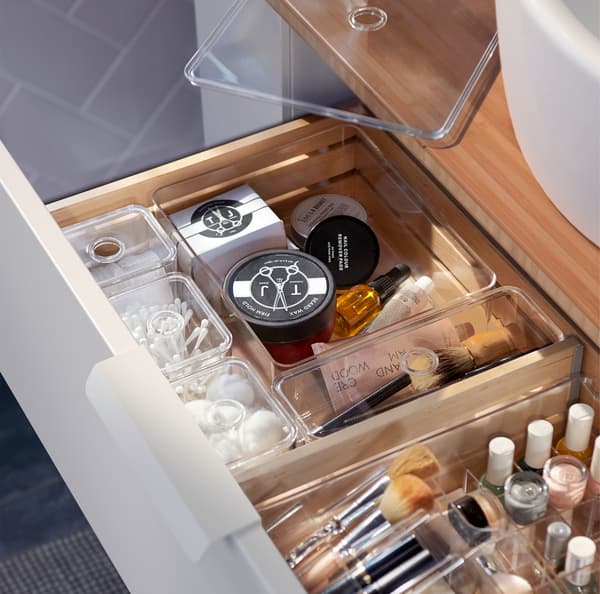 Unha combinación de caixas de almacenamento transparentes GODMORGON cheas de produtos de baño esenciais nun caixón de madeira.