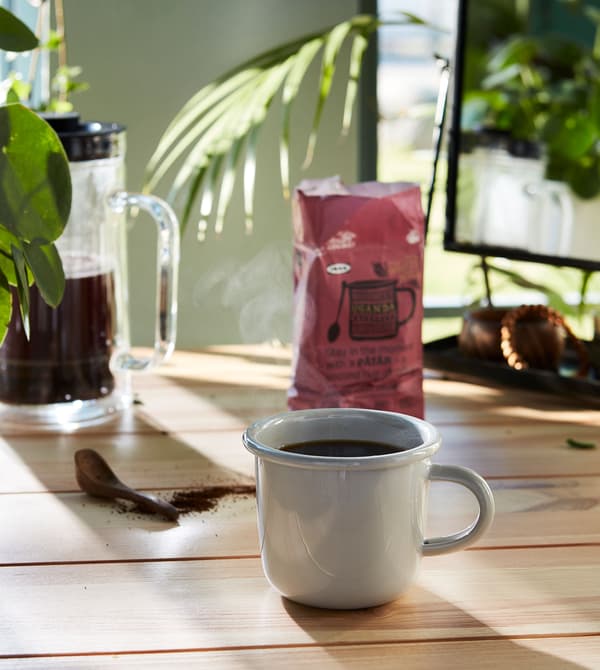 Unha cunca branca chea de café sobre unha superficie de madeira con café PÅTÅR e unha cafeteira ao fondo.