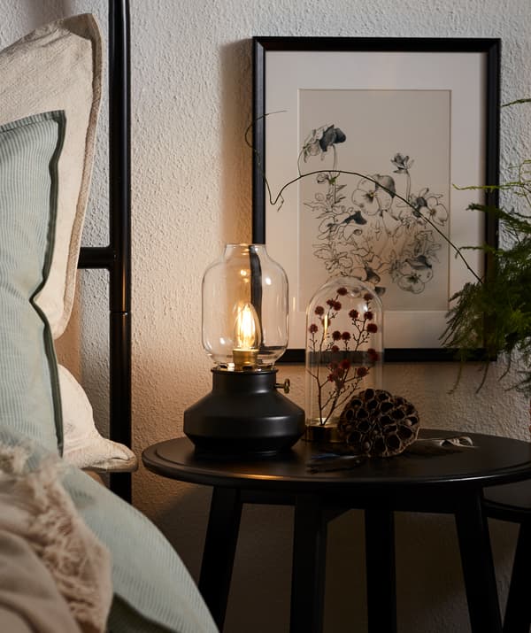 Unha mesa KRAGSTA colocada xunto á cama, decorada cunha planta seca dentro dunha campá de vidro e unha lámpada TÄRNABY cunha bombilla LED regulable.