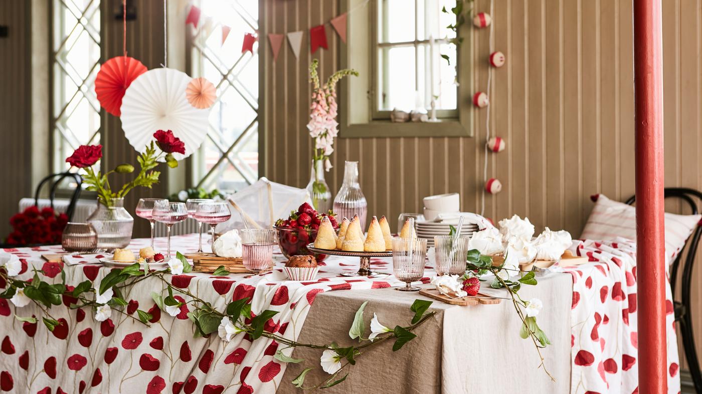 Unha mesa preparada para unha festa con distintos elementos da colección de tempada ANLEDNING, como vasos, flores e adornos brancos e vermellos pendurados da parede e do teito.