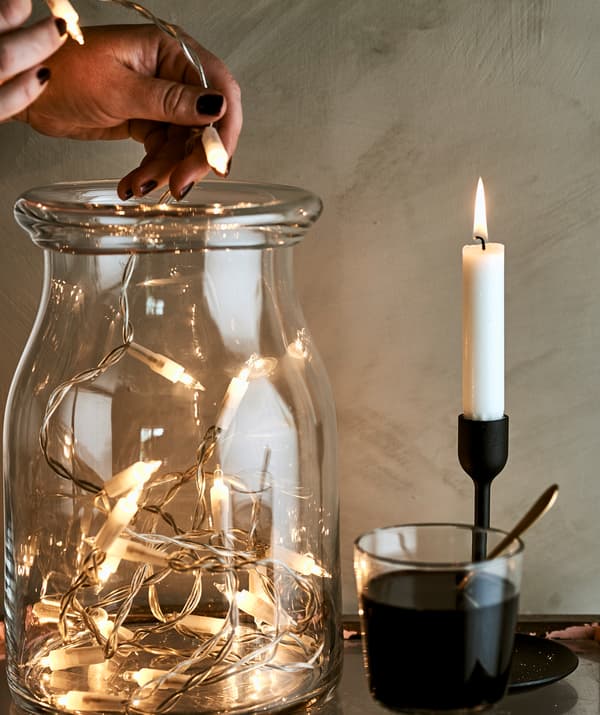 Unha muller enche un tarro con luces decorativas xunto a un vaso con viño e unha candea acesa nun portacandeas negro.