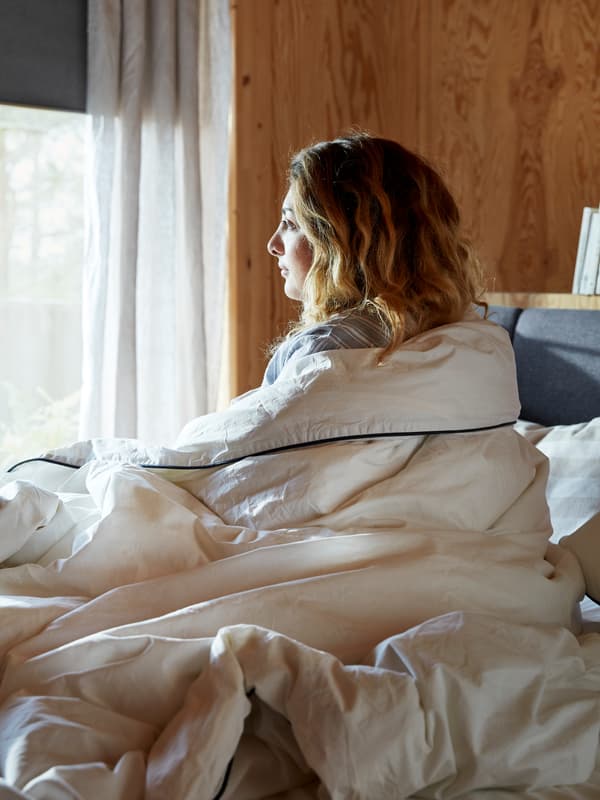 Unha muller na cama, en posición vertical e cuberta por un edredón de cor crema, contempla o exterior soleado pola ventá.