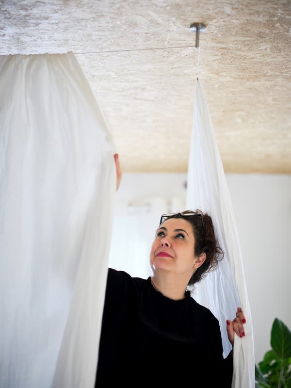 Unha muller pecha dúas cortinas de algodón brancas montadas no teito que serven de divisor de estancias. Nun recuncho hai unha planta grande.