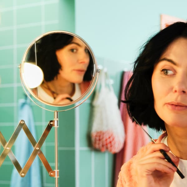 Unha muller suxeitando unhas tesoiras, ollando un espello FRÄCK orientable namentres corta o pelo nun cuarto de baño verde.