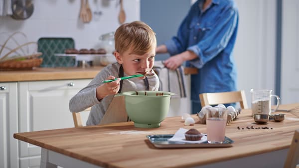 Unha persoa adulta observa a un neno nunha mesa PINNTORP en marrón claro mentres come masa para un pastel de chocolate dun cunco verde cunha espátula.