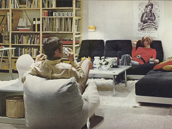 Unha sala de estar dos anos 1980 con dúas persoas sentada, con varios estantes IVAR ao fondo.
