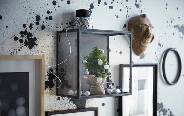 Unha vitrina IKEA BARKHYTTAN nunha parede, cunha árbore en miniatura con adornos, xunto con marcos de cadros.