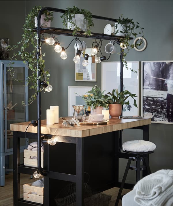 Un'isola cucina con rastrelliera VADHOLMA. Di fronte, uno sgabello da bar. Sul bancone ci sono piante in vaso, candele a led e un servizio da tè. Sulla rastrelliera ci sono luci decorative accese.