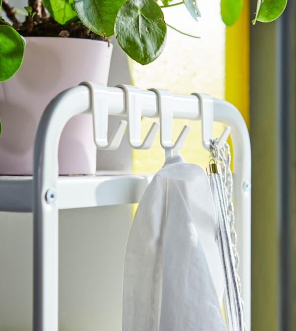 Uno scaffale a giorno bianco con ganci sul lato a cui sono appese collane e un indumento. Sopra, un vaso rosa con pianta.