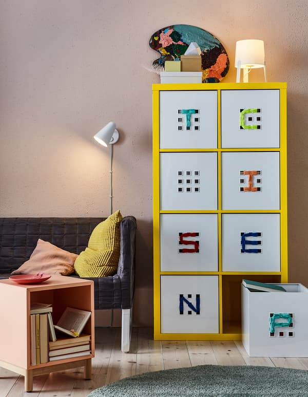 Uno scaffale giallo con contenitori LEKMAN decorati con un ricamo. A lato, un comodino rosa e un divanetto con cuscini colorati rosa e senape SVARTPOPPEL.