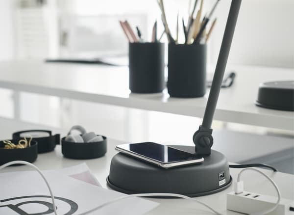 Uno smartphone in carica sulla base di una lampada da lavoro HEKTAR grigio scuro, su una scrivania bianca MITTZON.