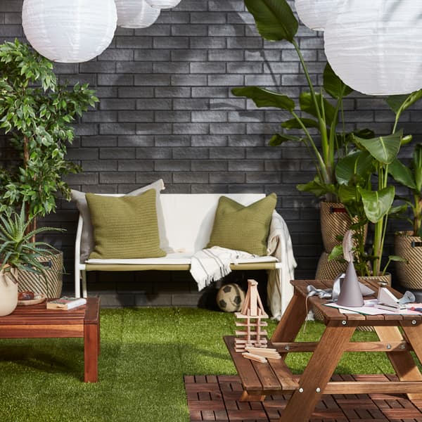Uno spazio esterno con delle accoglienti sedute, piante rigogliose, lampade a sospensione a LED a energia solare contro una parete in mattoni - IKEA