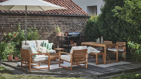 Uno spazio esterno soleggiato e con pedana in legno vicino a un muro in mattoni, arredato con il tavolo e le sedute NÄMMARÖ e un barbecue GRILLSKÄR - IKEA.