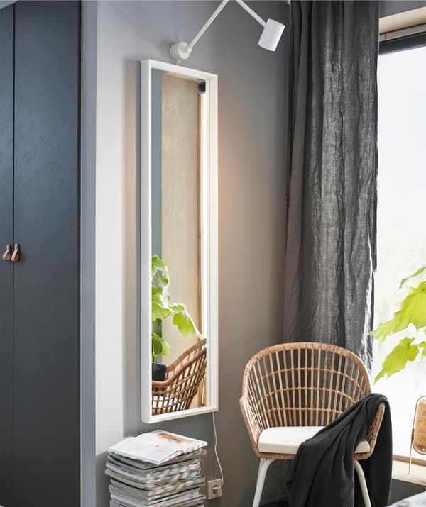 Uno specchio a figura intera accanto a una pila di riviste e una sedia in rattan sullo sfondo di una parete grigia.