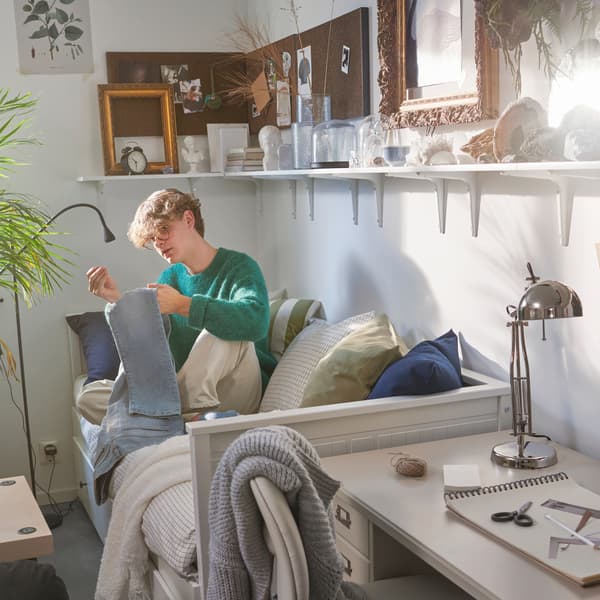 Uno studente seduto su un letto divano HEMNES mentre cuce un paio di jeans. Sulla parete ci sono delle mensole con una sveglia, cornici e oggetti - IKEA.