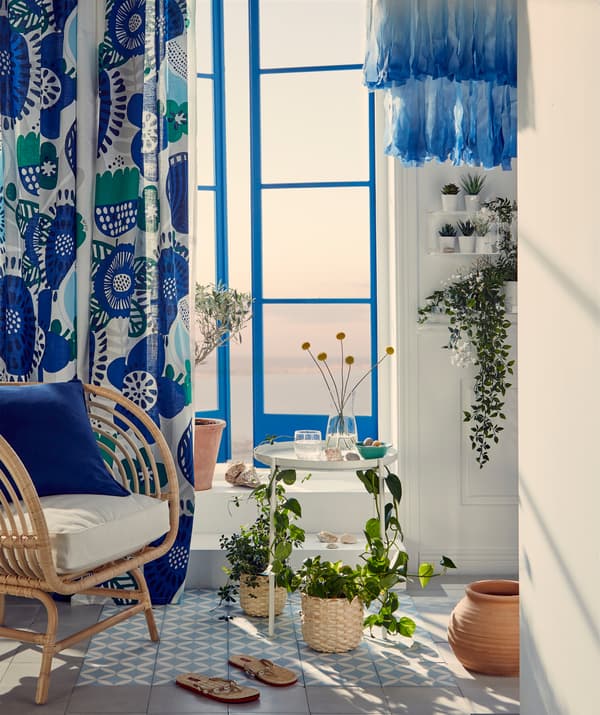 Un'oasi di verde davanti alla portafinestra di un balcone, una poltrona in vimini e piante che decorano mensole e un tavolino vassoio.