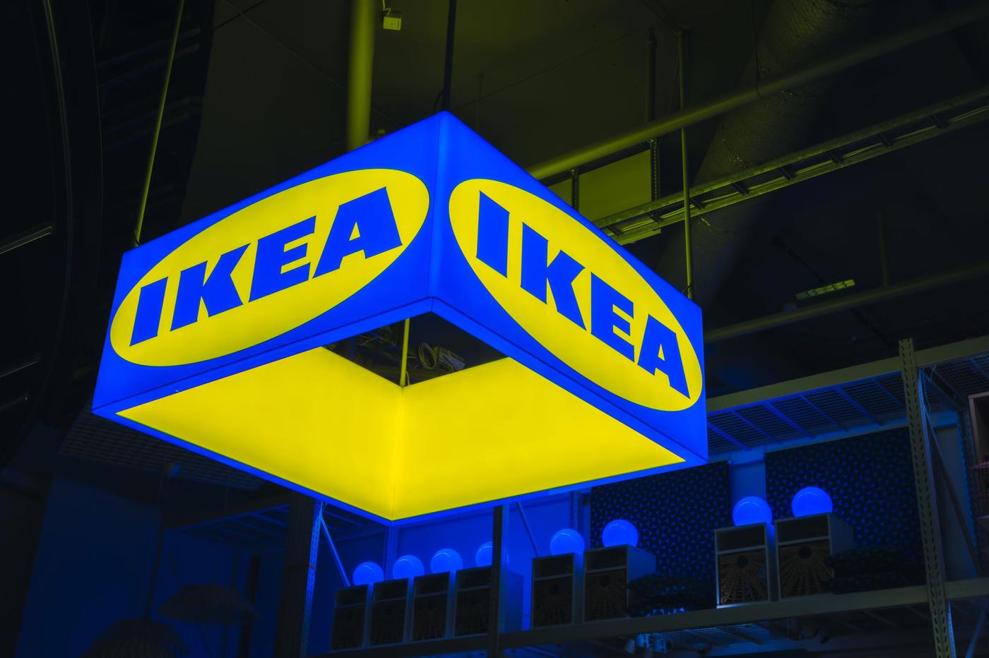Upplyst fyrkantig IKEA‑skylt i blått och gult hängande från taket inne i IKEA‑varuhuset i Mall of Scandinavia, med hyllor och belysning synliga i bakgrunden.