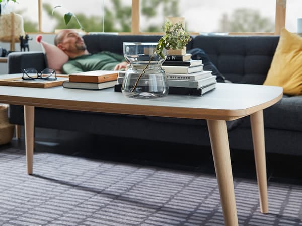 Uprostred je konferenčný stolík OMTÄNKSAM s vázou OMTÄNKSAM, knihami a okuliarmi navrchu. Muž odpočíva na pohovke v pozadí.