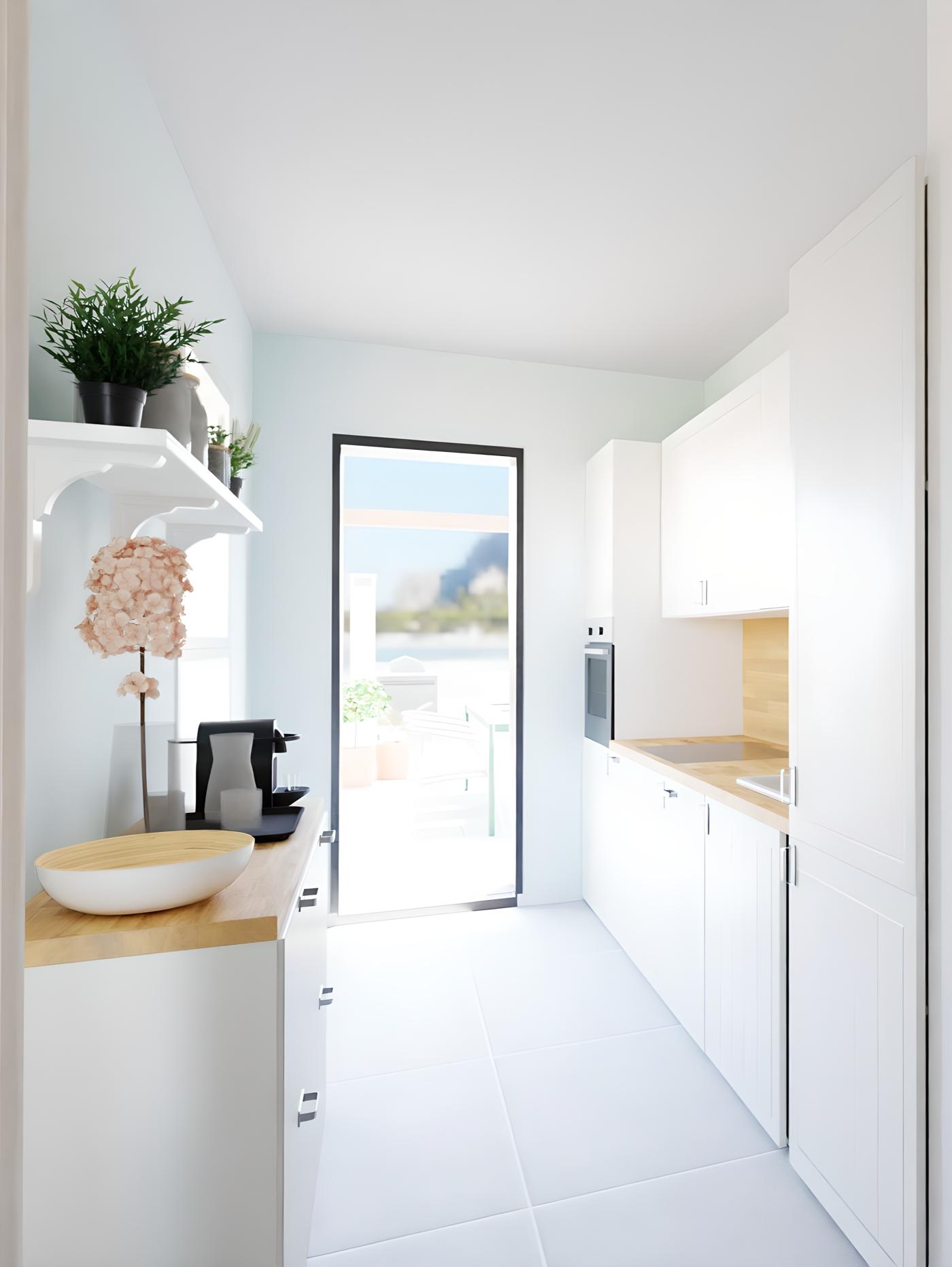 Uska, svijetla kuhinja s bijelim ormarićima, drvenim pločama, aparatom za kavu, zdjelom i cvijetom; kroz staklena vrata vidi se terasa s biljkama.