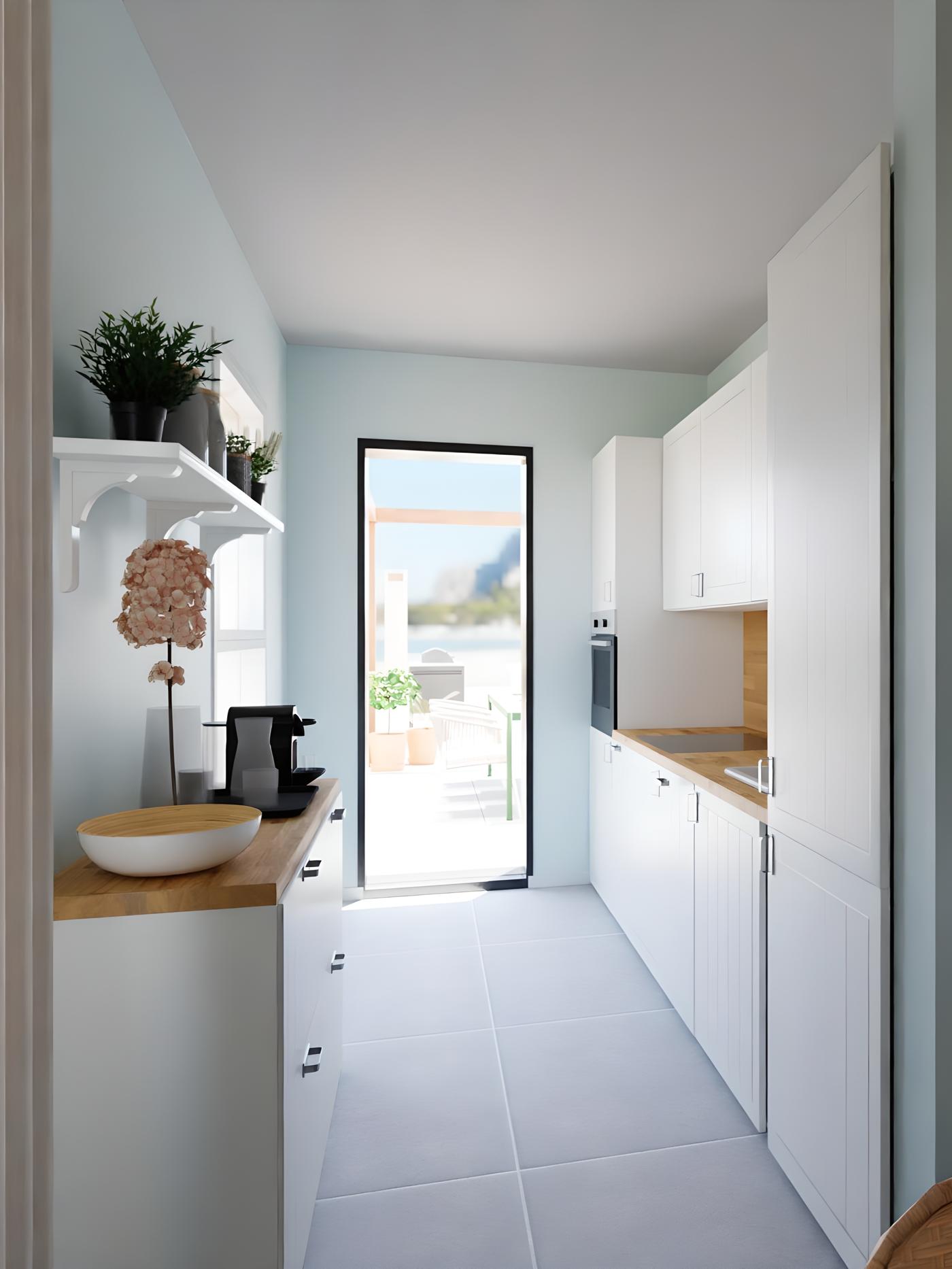 Uska, svijetla kuhinja s bijelim ormarićima, drvenim pločama, aparatom za kavu, zdjelom i cvijetom; kroz staklena vrata vidi se terasa s biljkama.